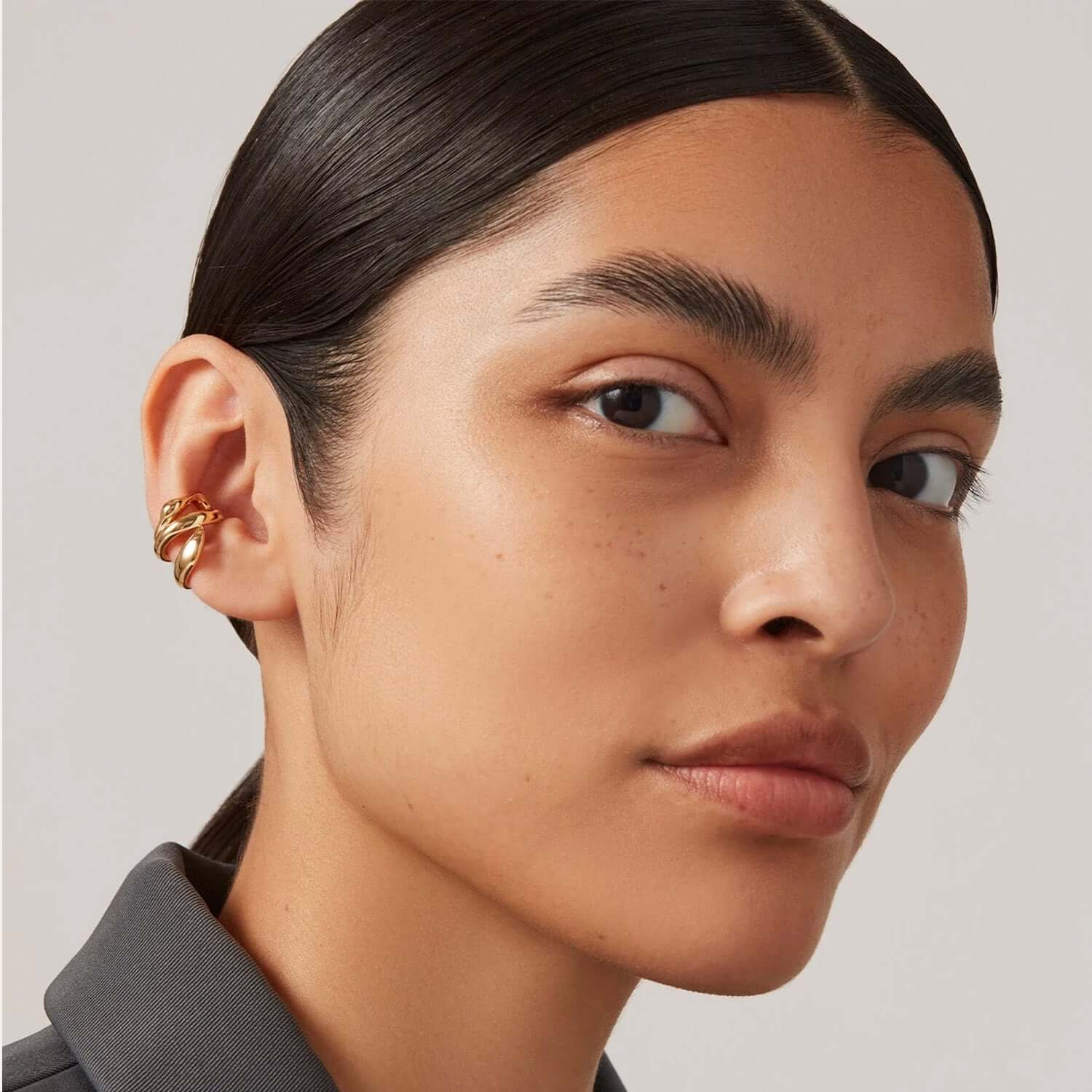 A model wearing the Earcuff Flow, a sculptural gold ear cuff that wraps the cartilage with liquid movement.