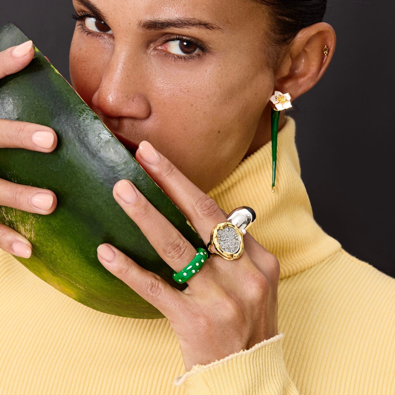Model showcasing Ring Coiné with a green pearl accent, holding a watermelon, in a stylish yellow sweater.