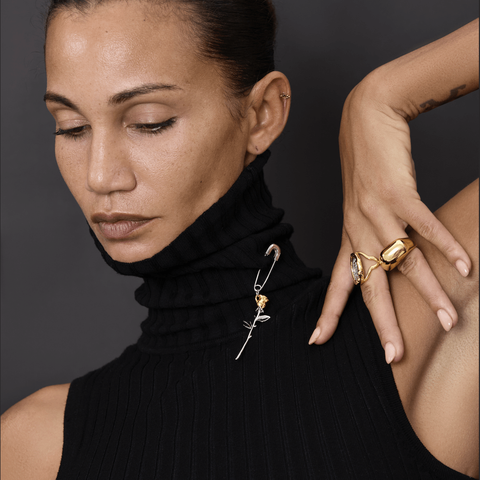 A close-up of a woman wearing Earrings Bloom, featuring a gold rose and safety pin design on a black turtleneck.